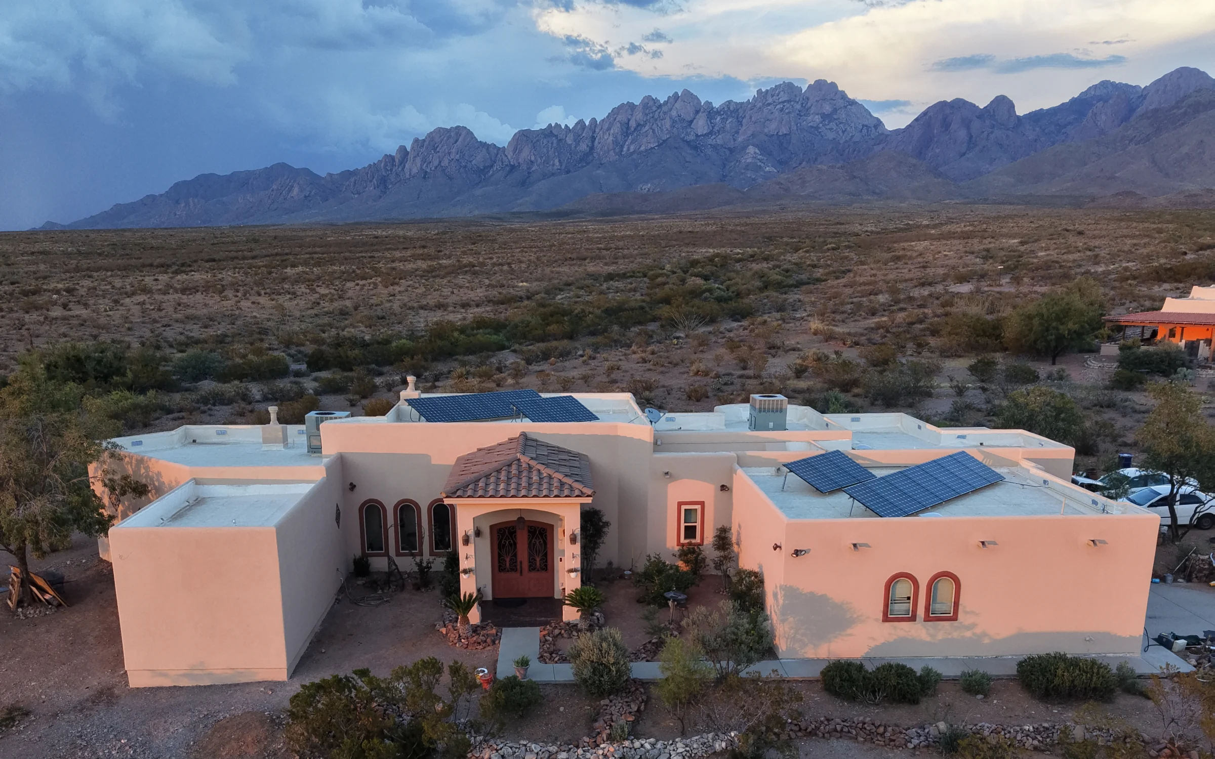 Pale stucco home glowing in warm evening light beneath a dramatic mountain ridgeline and cloudy sky.