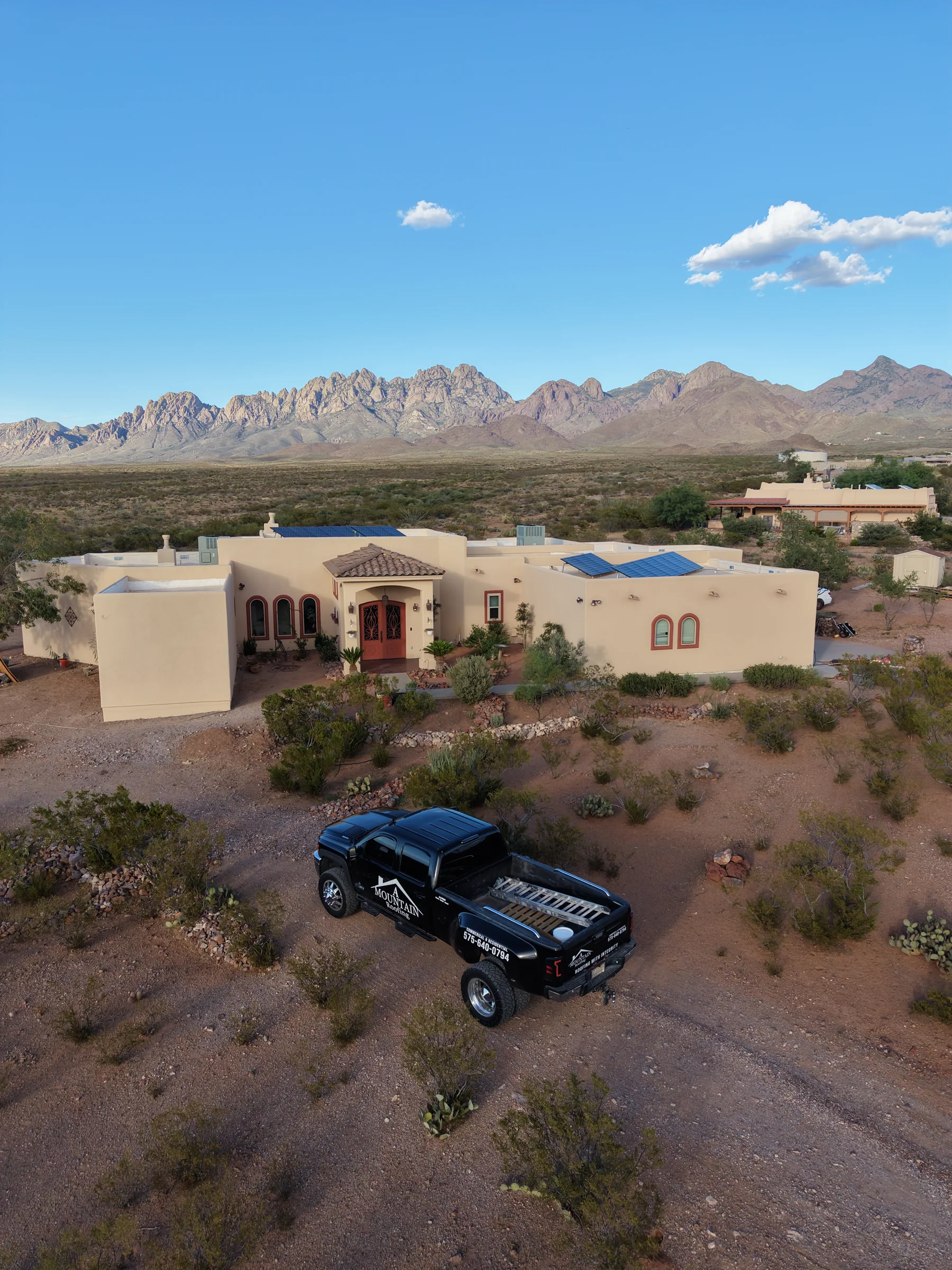 Pale stucco home with an arched entrance, solar panels, desert landscaping and a black pickup beneath a mountain backdrop.