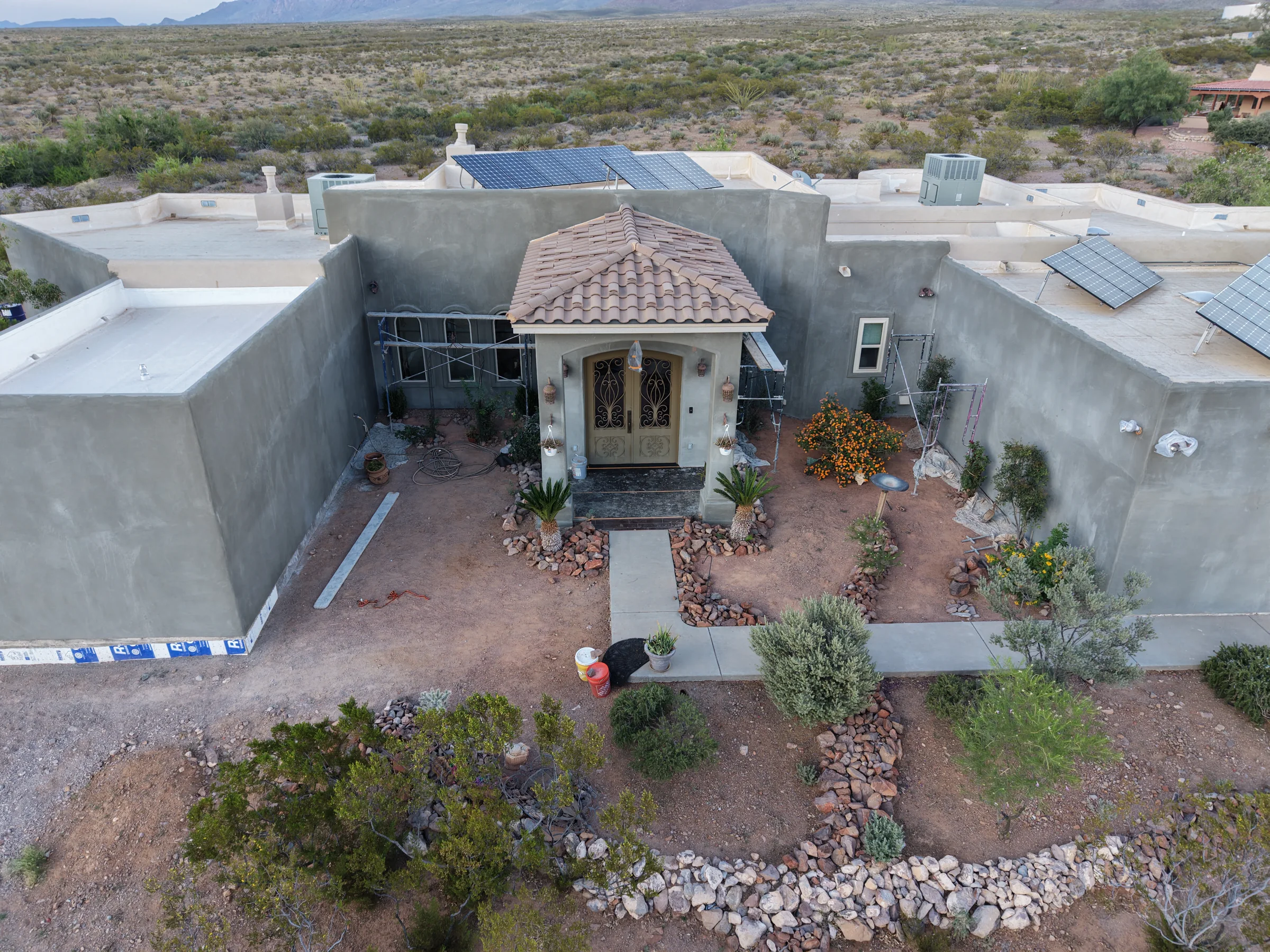 Gray stucco walls and scaffolding around an ornate courtyard entrance, with solar panels and desert landscaping.