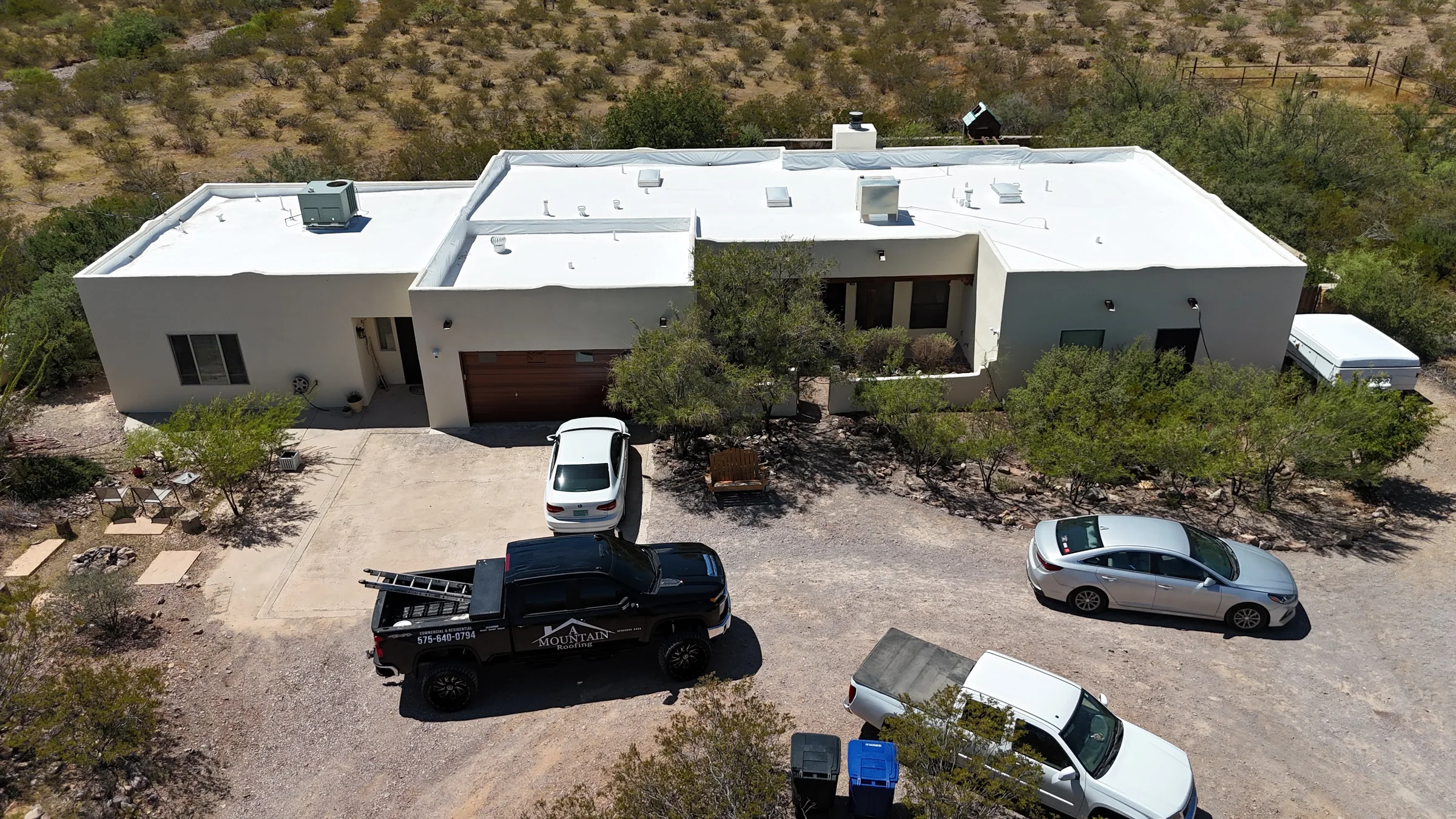 Aerial view of a pale stucco home with a garage, white flat roof, trees and vehicles in the driveway.