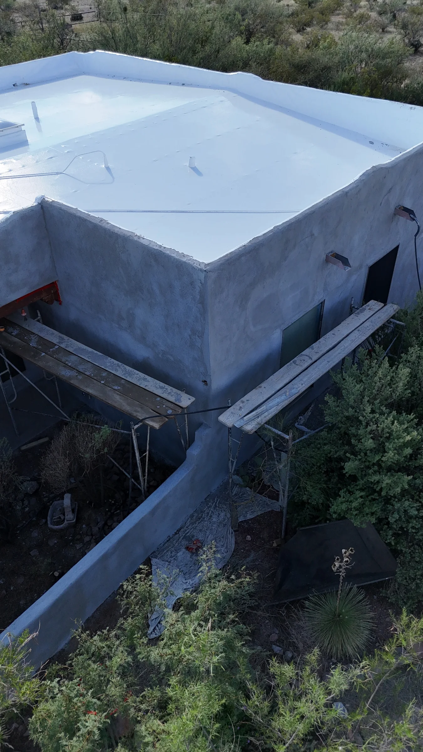 Gray stucco walls beneath a white flat roof, with scaffold boards along the house and garden wall.