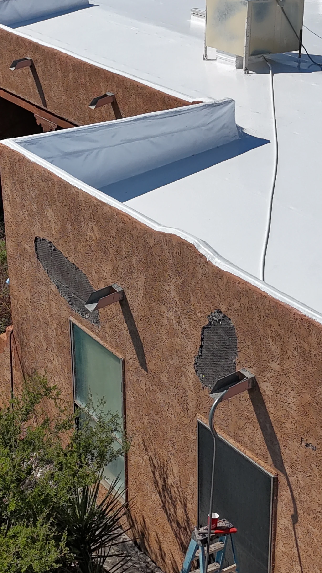 Textured brown stucco wall with gray repair areas near projecting roof drainage outlets below a white flat roof.