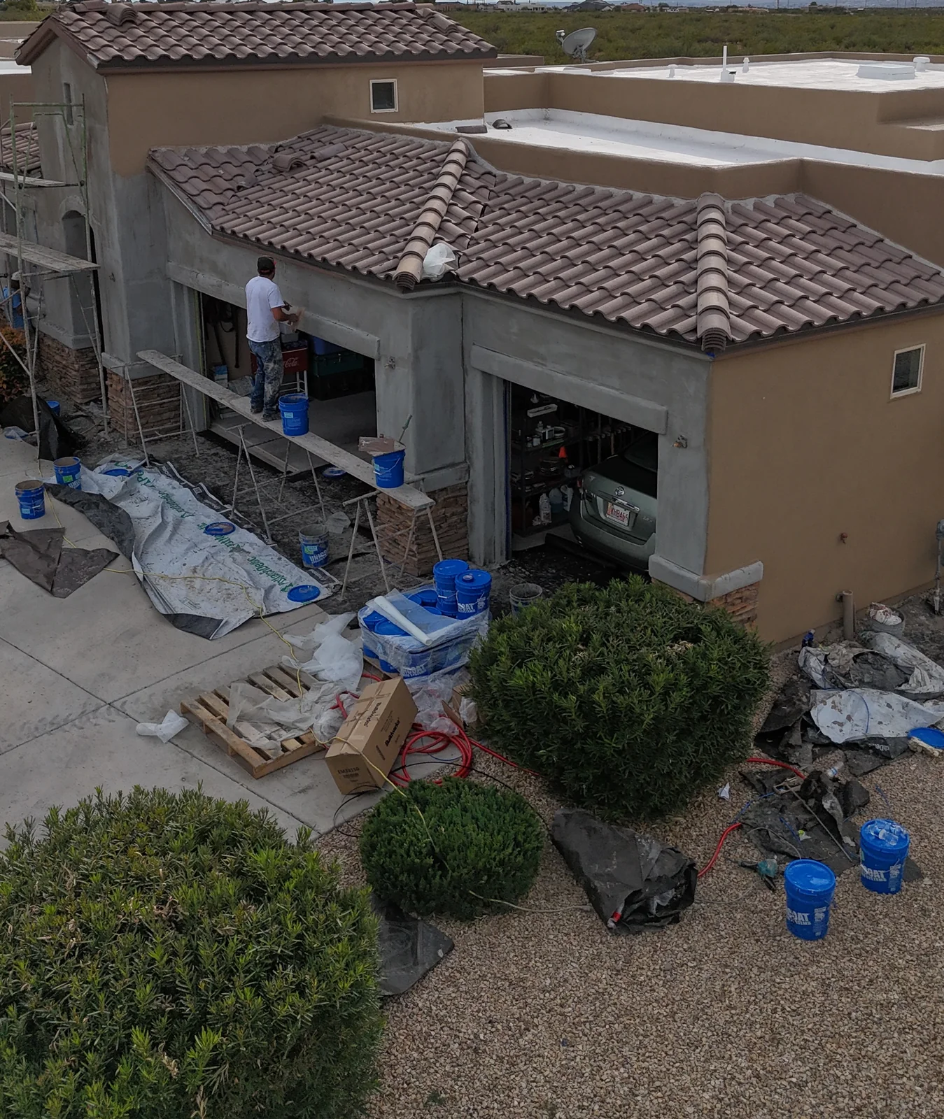 Worker on scaffolding beside unfinished gray stucco around the garage openings of a tan home.